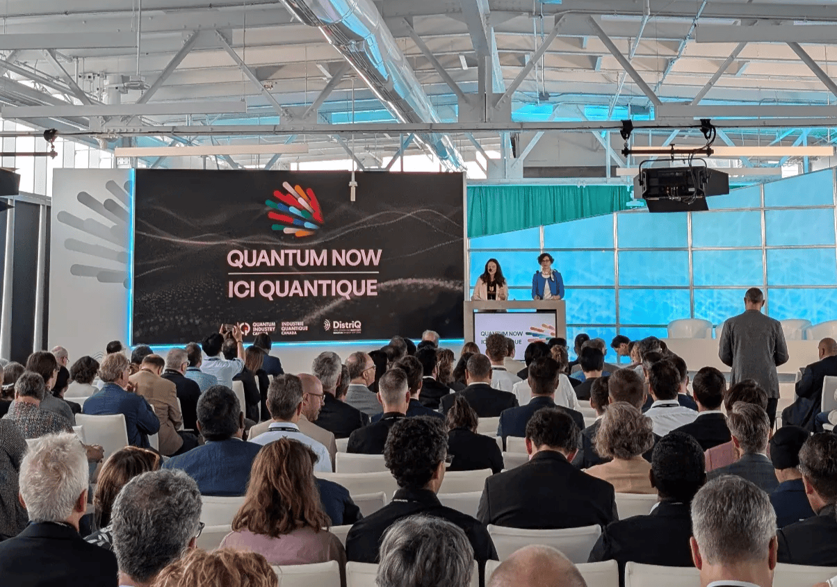 Speakers at the Quantum Now 2025 opening session standing behind a podium, with the event’s vibrant logo displayed on a large screen behind them.