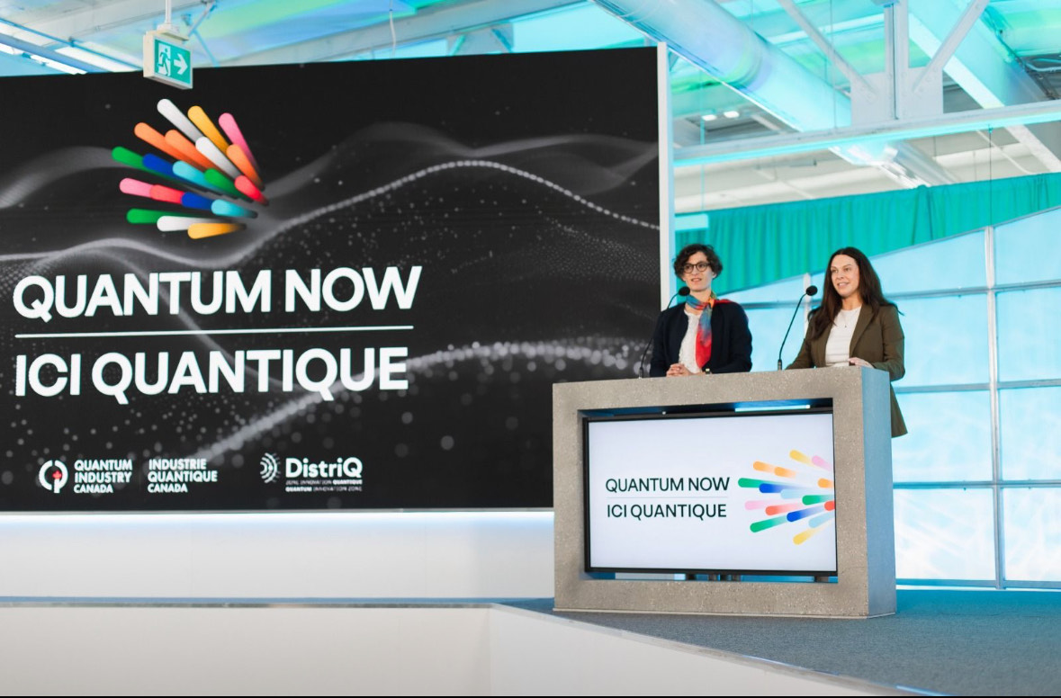 Speakers at the Quantum Now 2025 opening session standing behind a podium, with the event’s vibrant logo displayed on a large screen behind them.