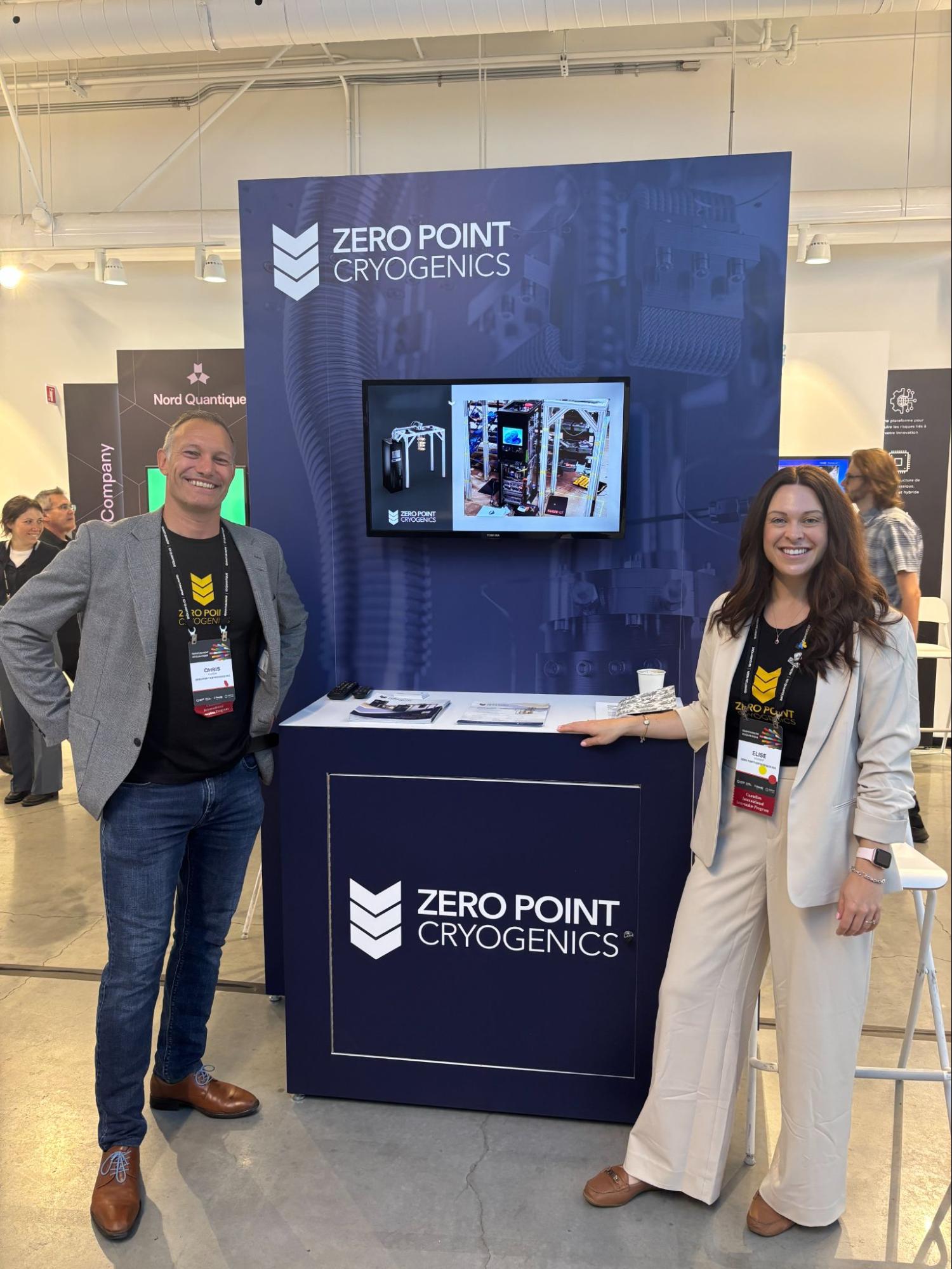 Zero Point Cryogenics team members, Chris and Elise, standing at their branded booth at Quantum Now 2025 in Montréal, with a display showcasing cryogenic technology on the screen.
